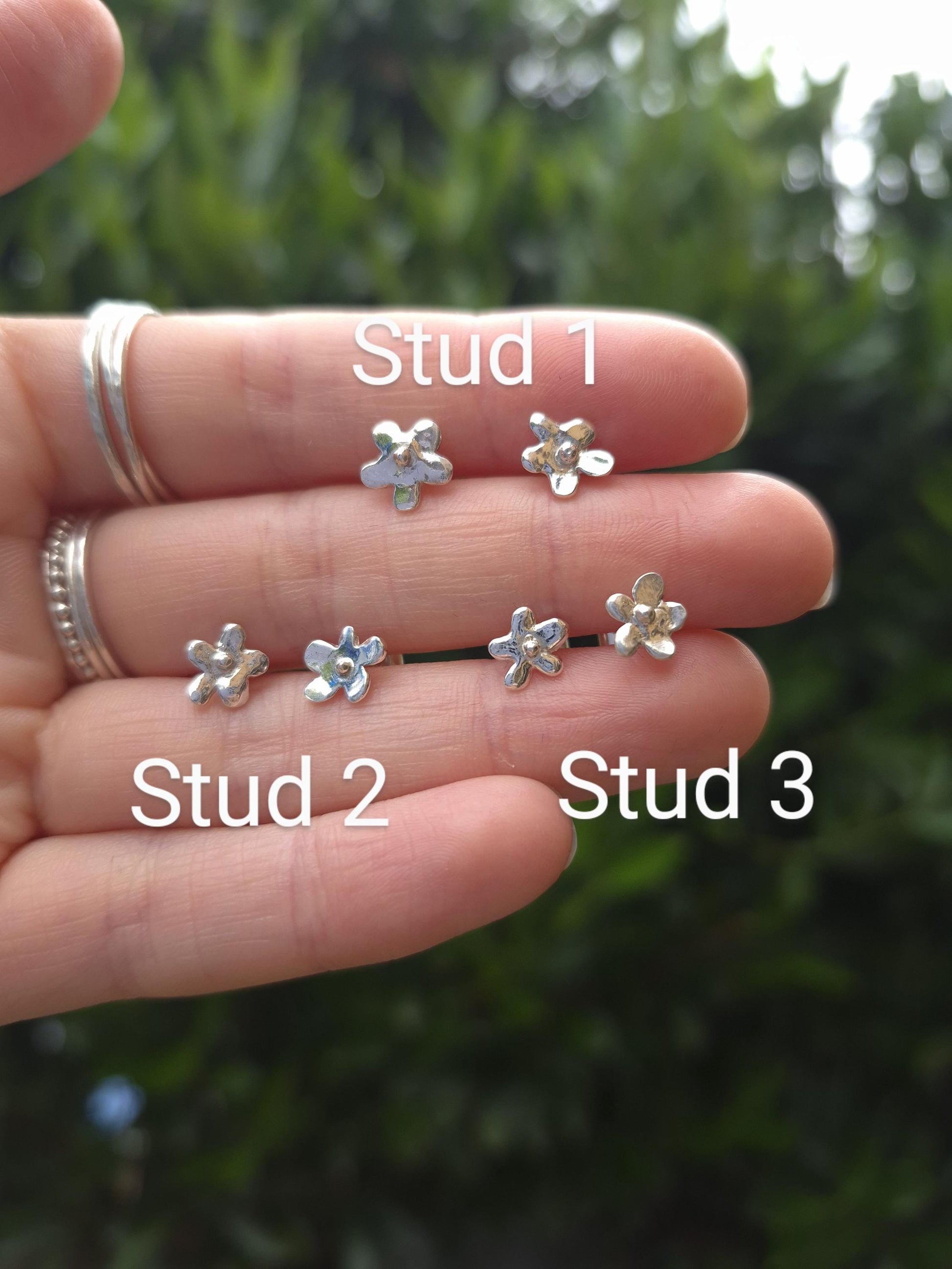 Three pairs of silver flower earrings on a hand with a blurred green background