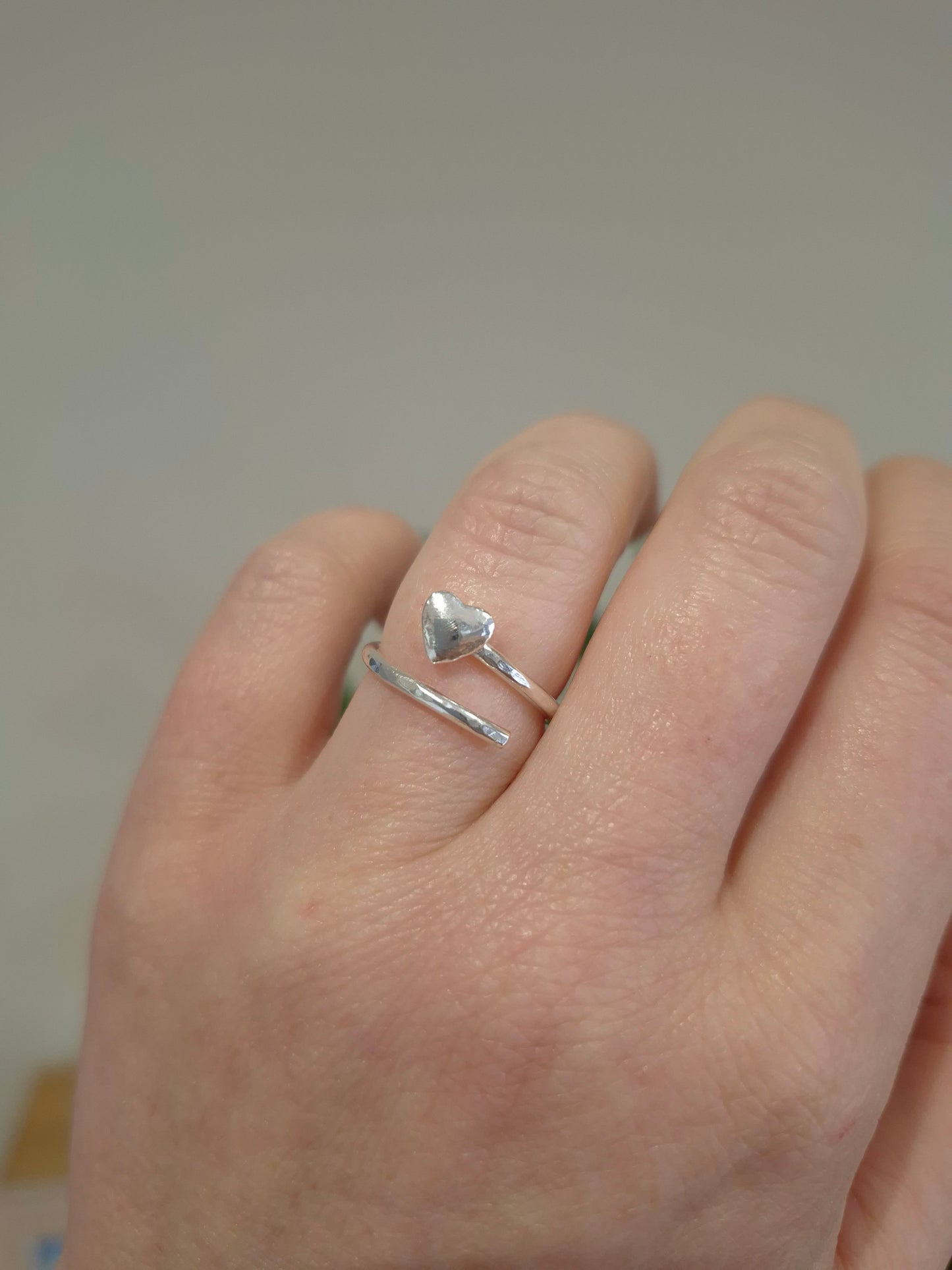 Hand wearing a silver ring with heart design that wraps around finger on a neutral background