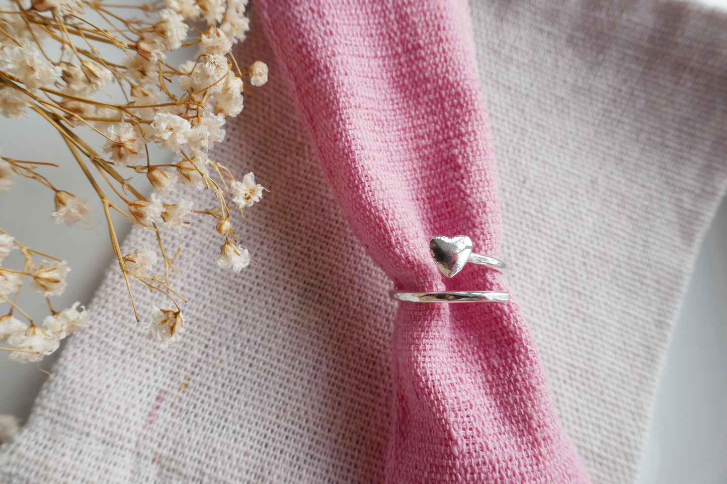 Silver ring with a heart design on a pink fabric background with dried flowers.