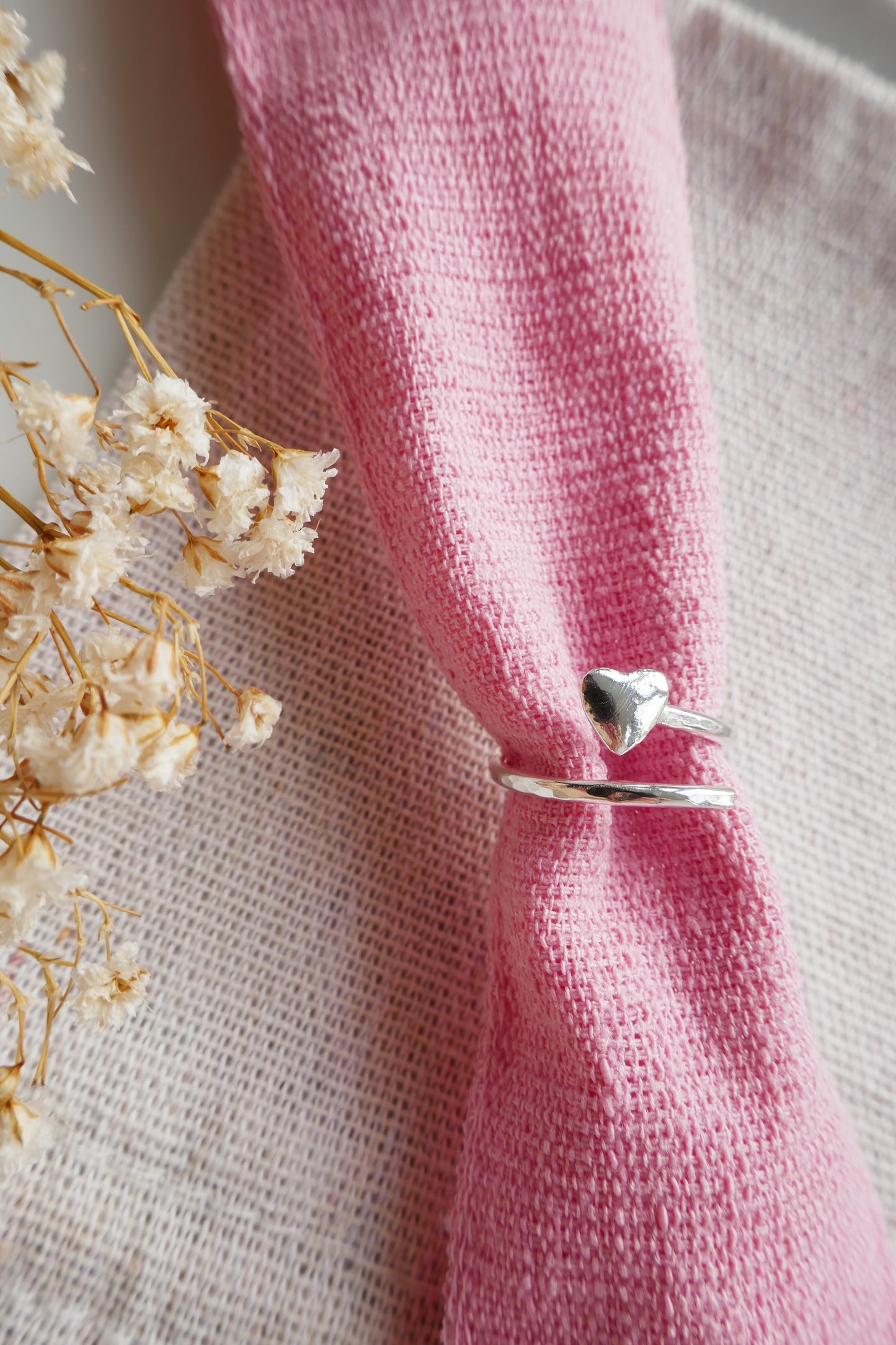 Silver ring with heart detail on a pink knitted fabric with dried flowers in the background