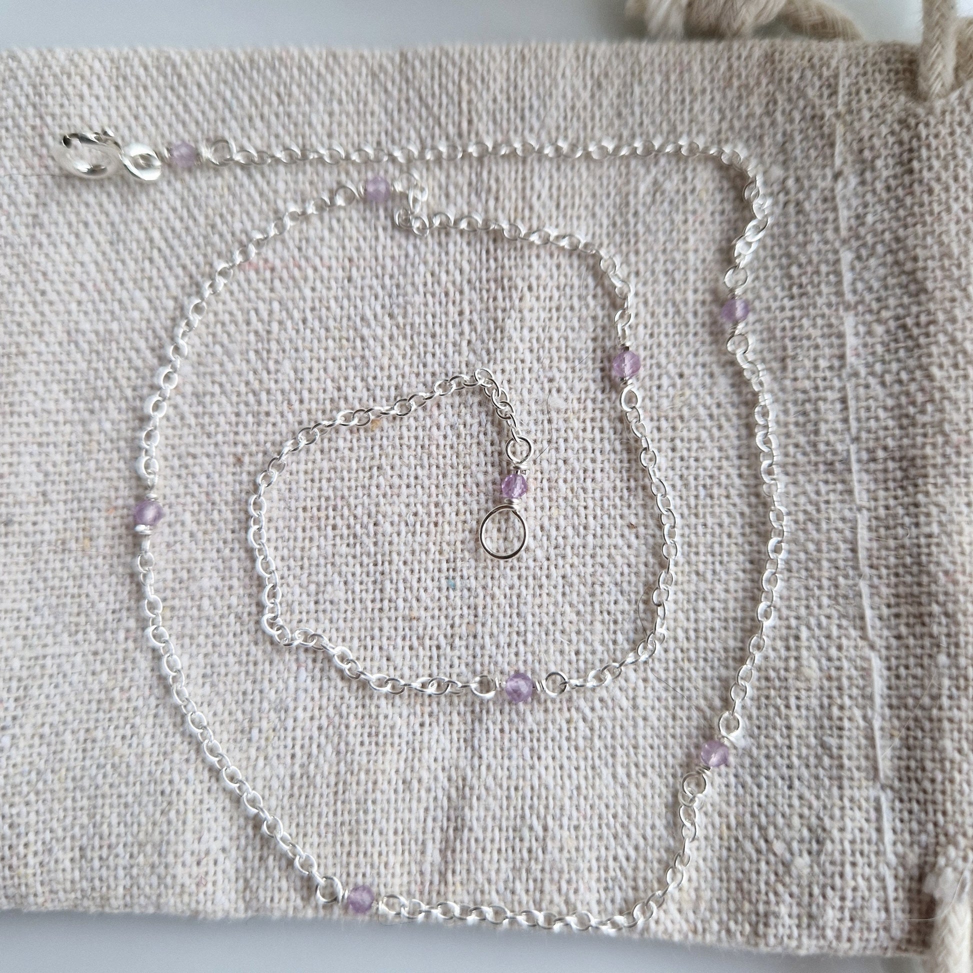a delicate silver chain holds nine tiny light purple amethyst beads dotted along it. The necklace is placed upon a pale fabric on a white background.