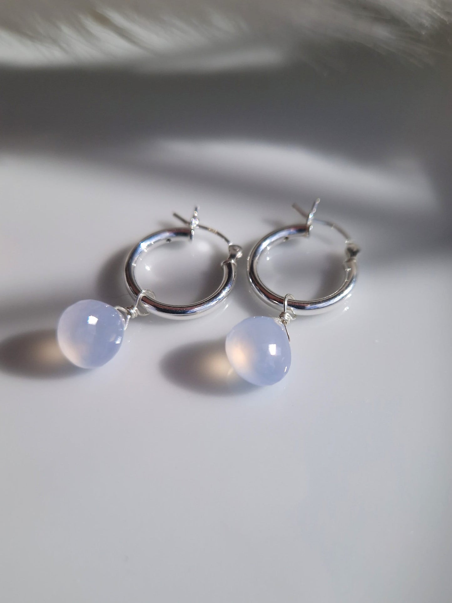 chalcedony gemstone and sterling silver hoop earrings

A white background with two small silver hoop earrings with one wire wrapped blue gemstone dropping from each hoop. The gemstones are faceted and reflect a golden light from within the blue.